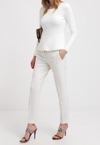 Vrouw in een witte textuur langemouwen top, witte op maat gemaakte broek en slangenhuidpatroon hoge hakken, staand met één hand in de zak.