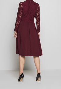 Robe bordeaux avec un corsage en dentelle et des manches longues, dotée d'une jupe plissée et d'une fermeture éclair à l'arrière. Chaussures à talons hauts noires.