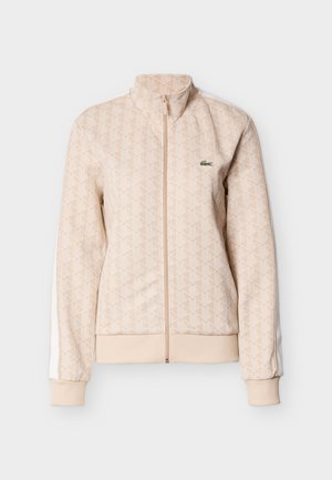 Bēša rāvējslēdzēja jaka ar ģeometrisku rakstu, ribota aproces un apakšmala, balti sānu svītras; uz krūtīm ir mazs zaļš Lacoste logo.