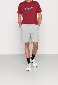 Červené bavlněné tričko s modrým logem Nike, šedé bavlněné šortky s tkaničkou, bílé tenisky, černé ponožky, stojící na neutrálním povrchu.