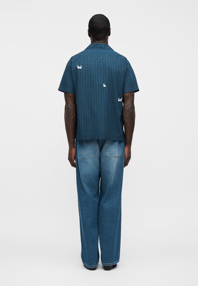 Πουκάμισο με κοντά μανίκια σε σκούρο πράσινο καρό με λευκές κεντητές λεπτομέρειες, συνδυασμένο με φαρδιά μπλε ξεβάμμένα τζιν, που φαίνεται από πίσω.