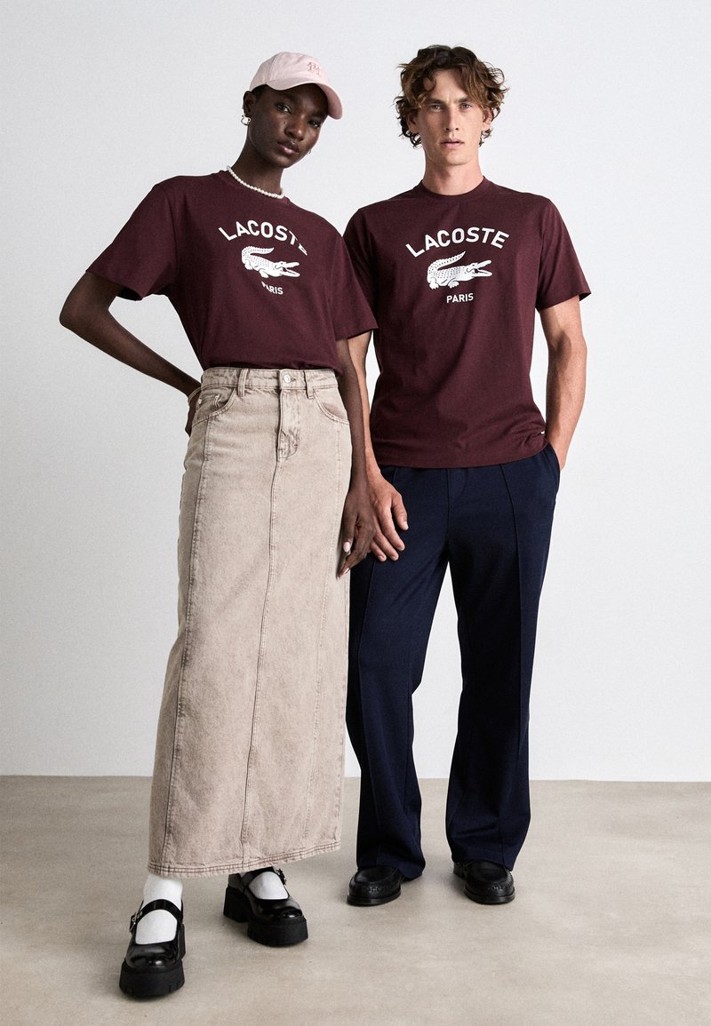 Camisetas de algodón marrón con el logo blanco de Lacoste, combinadas con una falda de mezclilla beige y pantalones azul marino. Zapatos de plataforma negros y una gorra rosa claro.