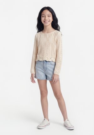 Usmievavá dievčina s dlhými čiernymi vlasmi, oblečená v béžovom pletenom svetri, svetlých rifľových šortkách a bielych teniskách, stojaca pred jednoduchým pozadím.