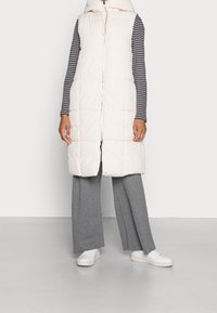 Lång, ärmlös puffer-väst i mjuk beige, med hög krage och dragkedja fram. Parat med grå vida byxor och vita sneakers.