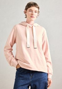 Persona che indossa un hoodie rosa chiaro con ampi lacci crema e jeans blu, in piedi con una mano in tasca su uno sfondo semplice.