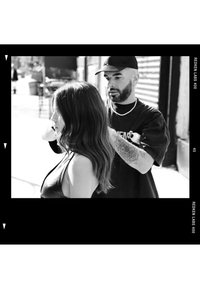 Homme coiffant les longs cheveux d'une femme en extérieur sur une photo en noir et blanc avec un arrière-plan urbain et un bras tatoué visible.