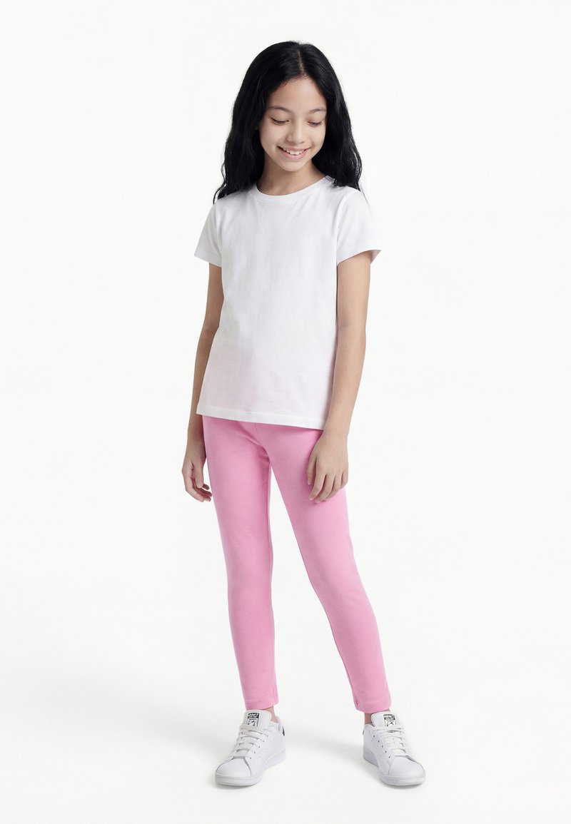 Κορίτσι με μακριά μαύρα μαλλιά που φοράει λευκό μπλουζάκι, ροζ κολάν και λευκά αθλητικά παπούτσια, στέκεται και χαμογελά με το κεφάλι ελαφρώς γερμένο προς τα κάτω.