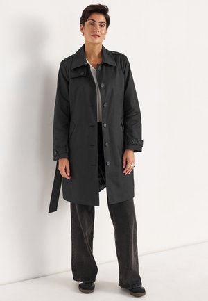 Woman standing in black buttoned trench coat, white top, black wide-leg pants, and black shoes against a plain white wall.