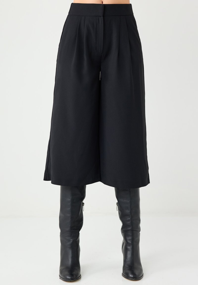 Czarne kuloty o wysokim stanie, z plisami, gładką teksturą i szerokimi nogawkami. Zestawione z czarnymi skórzanymi kozakiem do kolan.