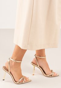 Sandalias de tacón alto doradas con tiras finas en el tobillo y tiras cruzadas. Diseño de punta cuadrada y acentos metálicos en un tacón de aguja. Textura suave.