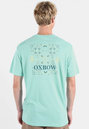 Homme aux cheveux blonds portant un t-shirt vert menthe avec des motifs géométriques et de palmiers, ainsi que le texte "OXBOW" dans le dos.