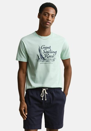 Mann trägt hellgrünes "Gant Sailing Race" T-Shirt und marineblaue Shorts mit Kordelzug, steht mit einer Hand in der Tasche vor einfarbigem Hintergrund.