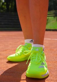 Nohy v neónovo zelenej športovej obuvi Adidas a bielych ponožkách Adidas stoja na hlinených tenisových kurtoch.