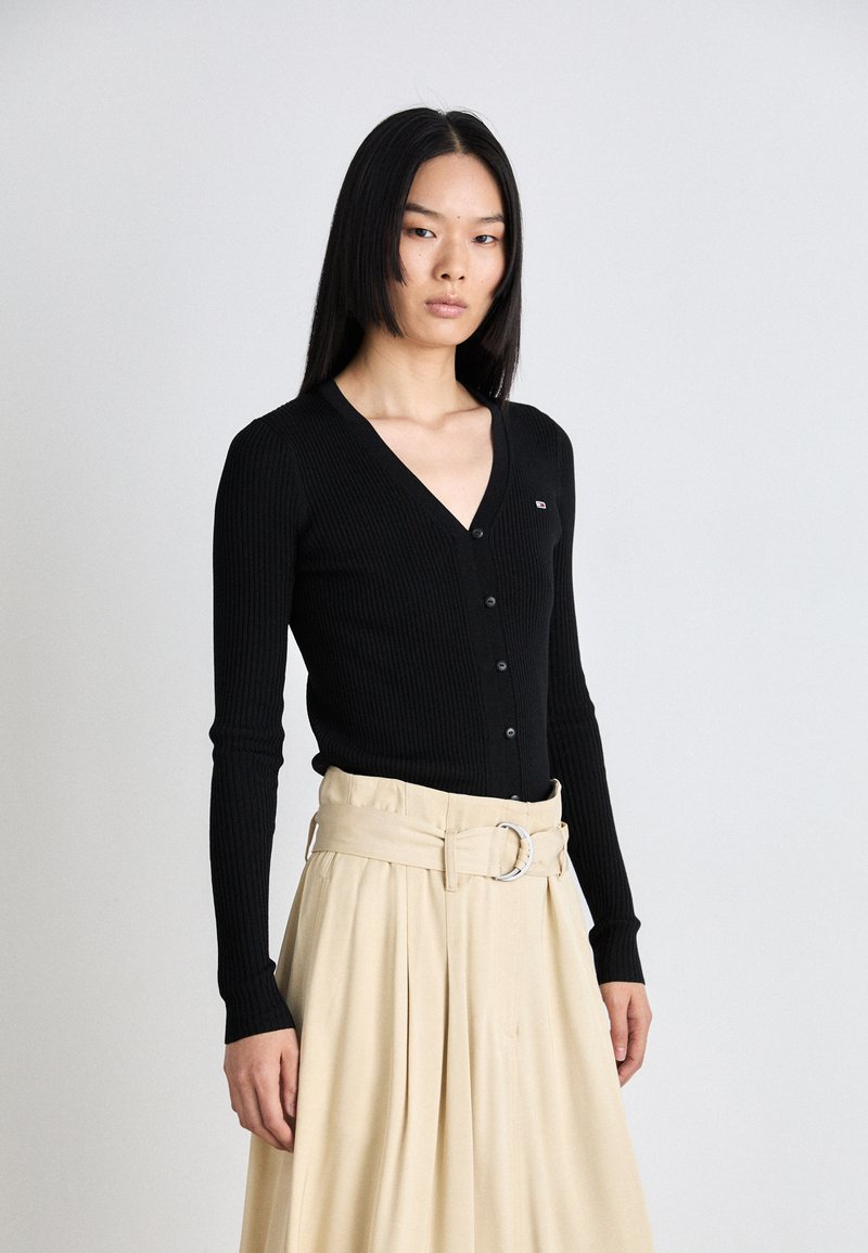 Melnā ribota kardigāna ar V veida kakla izgriezumu, garām piedurknēm un pogu aizdari. Saskaņots ar bēšu plīvētu svārku un platu jostu.