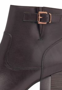 Stivaletto alla caviglia in pelle marrone con finitura opaca, dotato di una fibbia decorativa, accenti cuciti e un tacco in legno.