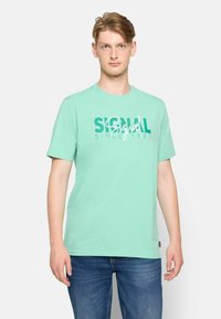 Camiseta de algodón verde menta con un gran estampado gráfico que presenta el texto "Signal" y "Desde 1967". Mangas cortas y cuello redondo.