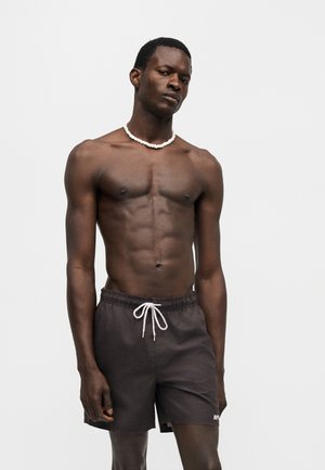 Homme noir athlétique portant un short de bain noir avec cordon et un collier blanc en perles, debout devant un fond clair uni.