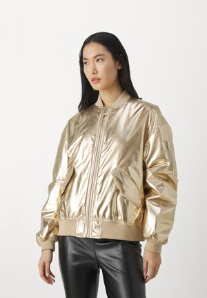 Goldene metallische Bomberjacke mit einem Reißverschluss, gerippten Bündchen und Saum sowie seitlichen Taschen. Getragen über schwarzen, engen Hosen.