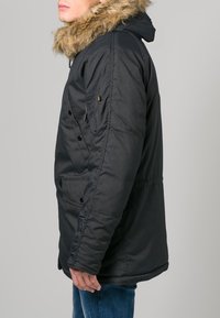 Parka nera isolata con cappuccio foderato in pelliccia sintetica, bottoni a pressione, tasche laterali e una vestibilità leggermente oversize. Caratterizzato da un tessuto liscio e opaco.