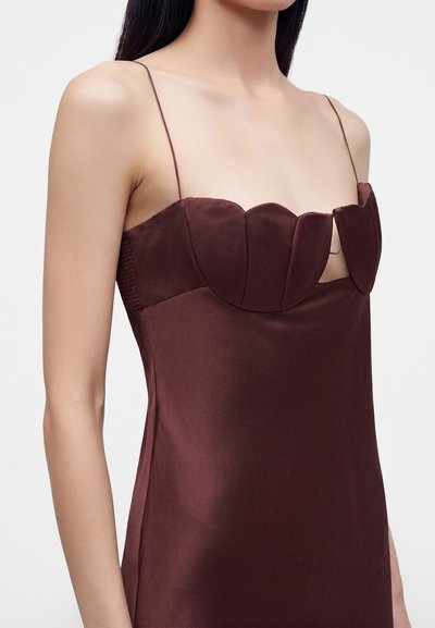 Femme portant une robe en satin marron avec de fines bretelles et un décolleté en cœur festonné unique sur un fond clair uni.