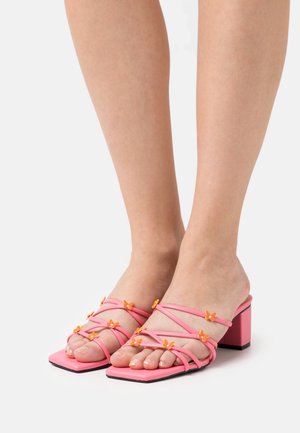 Pieds portant des sandales à talons roses avec des fines lanières ornées de petites décorations papillons orange sur un fond blanc.