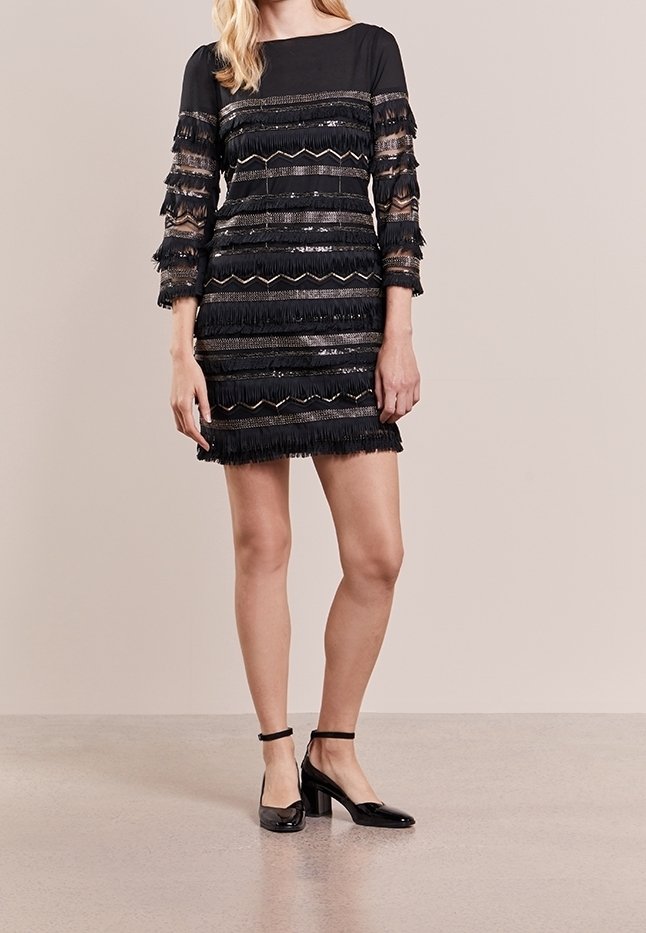 Robe noire avec des couches de franges horizontales, des accents métalliques et des manches longues. Associée à des talons en cuir verni noirs. Texture lisse, silhouette ajustée.