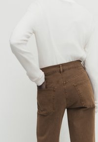 Bruna denimbyxor med hög midja, två bakfickor och synliga sömmar, kombinerade med en vit långärmad skjorta. Slät textur.