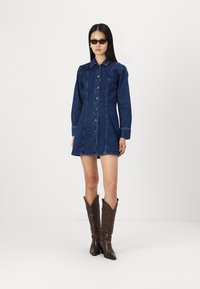 Denim srajčna obleka v temno modri barvi, z gumbnim zapiranjem spredaj, dolgimi rokavi in ovratnikom. Oblečena z rjavimi kavbojskimi škornji.