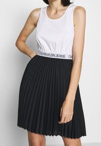 Vit linne med rundad halsringning, kombinerad med en svart veckad kjol med elastisk midja märkt "CALVIN KLEIN JEANS."