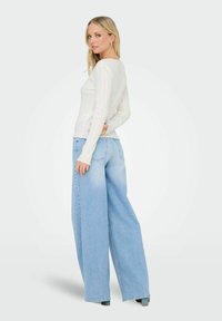 Witte geribbelde longsleeve top in combinatie met lichtblauwe wijde jeans. De top heeft een scalloped zoom; de jeans hebben achterzakken en een vervaagd uiterlijk.