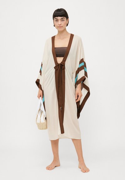 Robe style kimono crème avec des rayures brunes et turquoise, fermeture à nouer, manches larges et texture douce. Le mannequin tient un petit sac tissé.