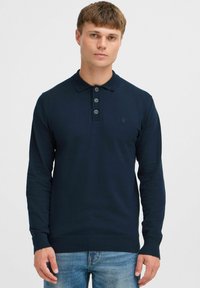 Polo à manches longues bleu marine avec col, trois boutons et poignets côtelés, associé à un jean bleu clair.