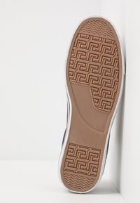 Semelle de basket en caoutchouc marron avec un motif géométrique et des accents blancs. Vue latérale sur un fond clair.