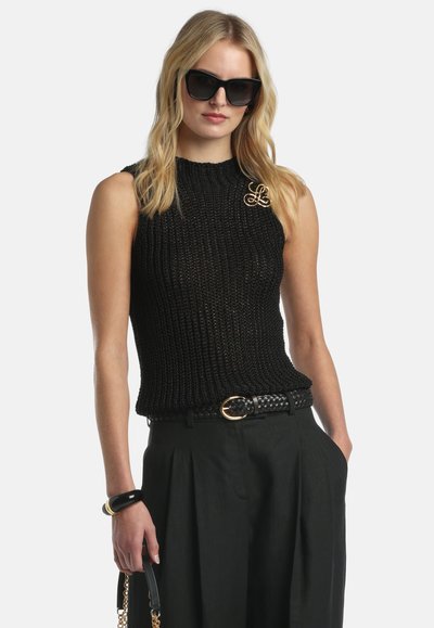 Plavolaska ženska oseba v črnem brezrokavnem pletenem vrhu, črnih sončnih očalih, črnih plisiranih hlačah, pletenem pasu, zapestnici in s črno torbico v roki.