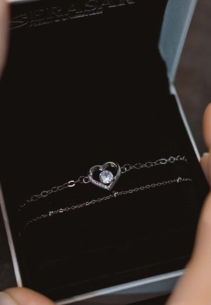 Silver bracelet with heart design featuring a central clear gem and small accents. Presented in a black velvet-lined box.