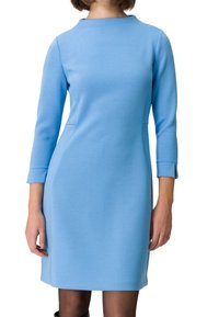 Femme portant une robe bleu clair ajustée, longueur genou, manches trois-quarts et col montant, debout devant un fond blanc.