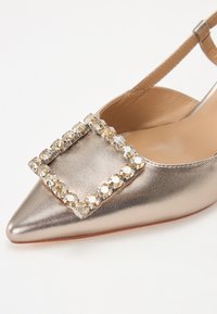 Fémes arany slingback cipő hegyes orral, nagy négyzet alakú strassz díszítéssel a lábfején és sima bőrfelülettel.