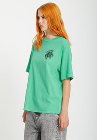 Zielony bawełniany t-shirt o luźnym kroju, z krótkim rękawem i czarną grafiką lamparta na piersi. Szare wzorzyste spodnie dopełniają całość.