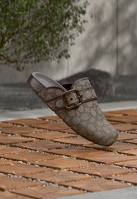 Brun mønstret slip-on sko med bred tåboks, struktureret design og guldfarvet metalspænde på en tekstureret overflade.