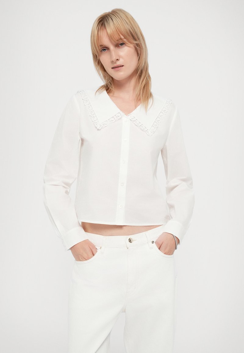 Weißes kurzes Hemd mit großem, mit Spitze besetztem Kragen, langen Ärmeln und Knopfleiste; kombiniert mit weißen Hosen; glattes, leichtes Material.