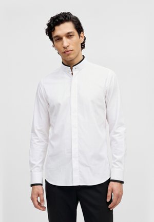 Un homme porte une chemise blanche à manches longues, bordée de noir au col et aux poignets, sur un fond clair uni.