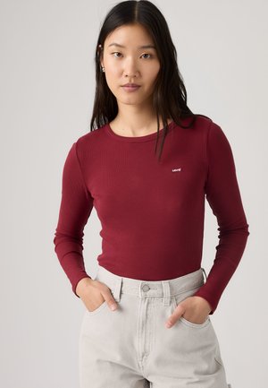 Femme aux longs cheveux foncés portant un haut ajusté bordeaux à manches longues et un pantalon taille haute gris clair, mains dans les poches, fond neutre.