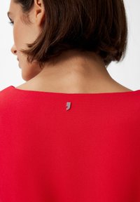 Rotes Oberteil aus glattem Stoff, mit weitem Ausschnitt und einem kleinen metallischen Akzent am Rücken. Einfaches, modernes Design.