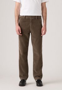 Pantaloni in corduroy marrone con vestibilità dritta, caratterizzati da una sottile trama a coste e una chiusura frontale con bottone, abbinati a scarpe nere.