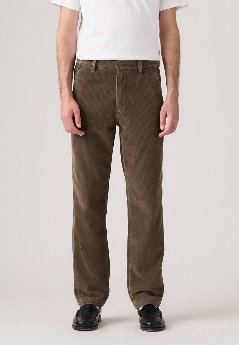 Pantaloni in corduroy marrone con vestibilità dritta, caratterizzati da una sottile trama a coste e una chiusura frontale con bottone, abbinati a scarpe nere.