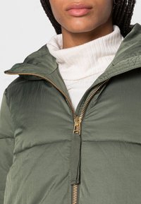 Grön puffjacka med texturerad yta, utrustad med en guldfärgad dragkedja och huva. Bärs över en vit ribbad polotröja.