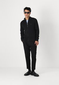 Svart ribbad zip-up tröja med hög krage, i kombination med svarta sluttande byxor och svarta skor. Modellen bär en grå t-shirt och solglasögon.