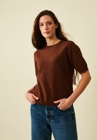 Ruskea lyhythihainen neule, jossa on pyöreä pääntie ja pehmeä rakenne, yhdistettynä sinisiin farkkuihin. Istuvuus on rento ja epämuodollinen.