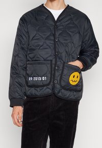 Veste matelassée noire avec un col en V, fermeture à bouton, deux poches avant, présentant un visage souriant jaune et des détails en texte blanc.
