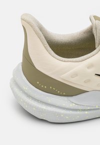 Zapatillas Nike Shield con una parte superior color crema y acentos en oliva, cuello texturizado y suela gris con motas amarillas.